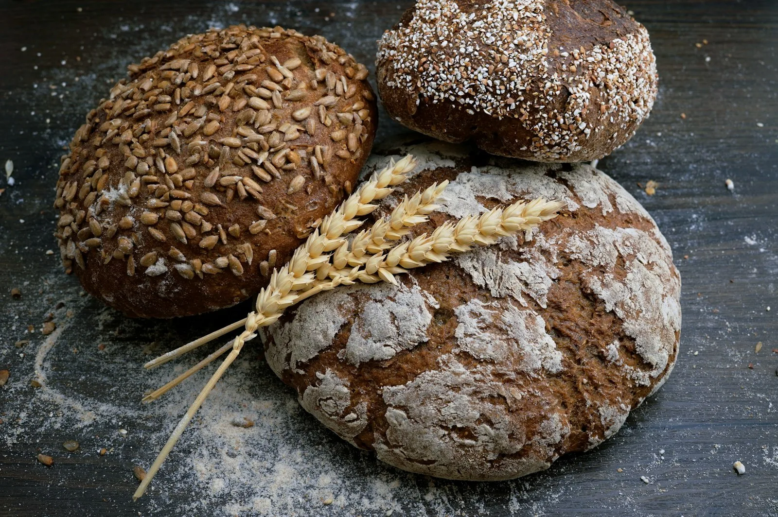 Three round rustic loaves on a floured dark bench, with wheat stalks laid across them