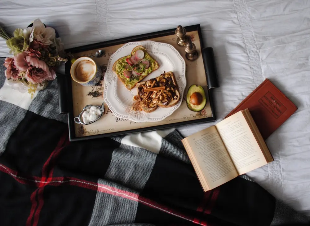 A breakfast tray with toast, coffee and a book on a made bed