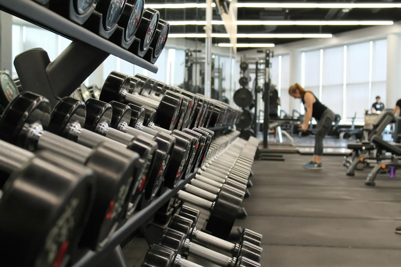 A long rack of dumbbells down one side of a bright gym floor, with lifting racks and benches at the far end