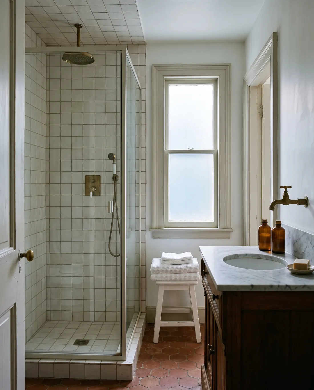 The guest bathroom: a walk-in rain shower behind glass, square off-white tile, and a marble-topped washstand with a brass tap