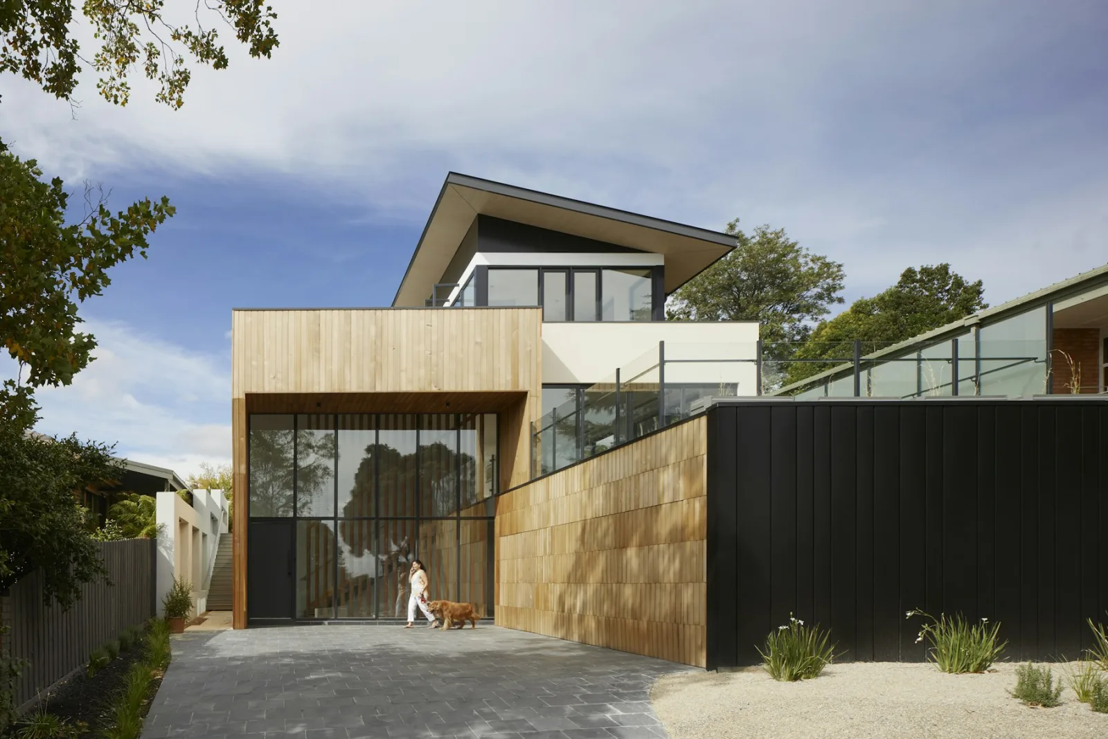 Two storey house in pale timber cladding beside a black metal wall, a wide angled roof over full height glazing