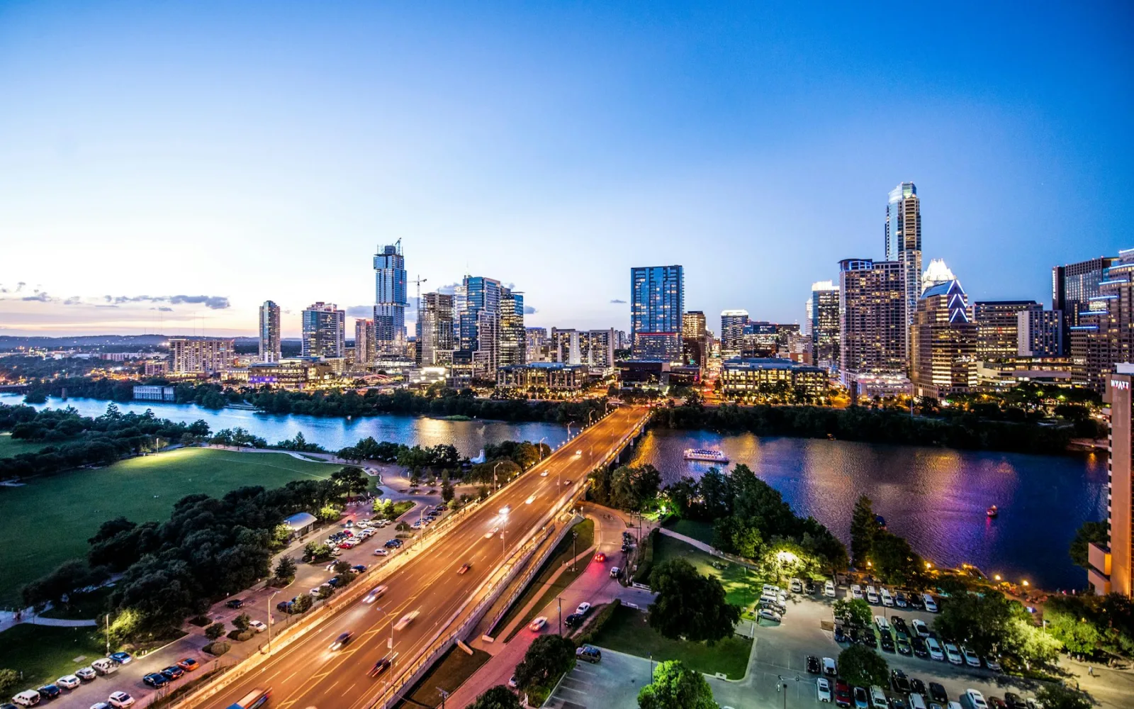 Downtown Austin at dusk, lit towers above Lady Bird Lake with traffic crossing the bridge and a park lawn on the near bank